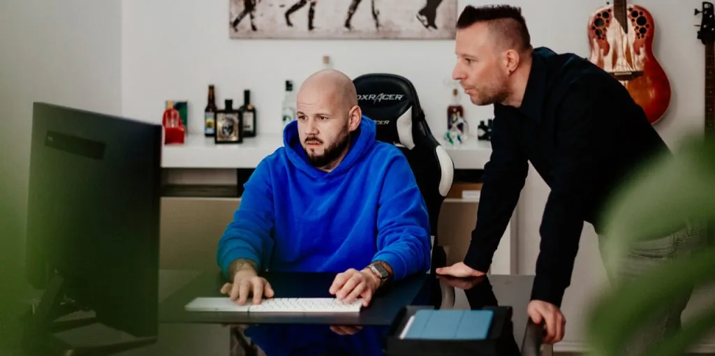 Die Gründer der Agentur wilde van rhee, Sebastian Winkler (links, im blauen Hoodie) und Alexander Boroš (rechts, im dunklen Hemd), arbeiten gemeinsam an einem Computer. Beide schauen konzentriert auf den Bildschirm. Im Hintergrund: Gitarren, Kunst und Spirituosenflaschen – ein kreatives, persönliches Büroambiente.