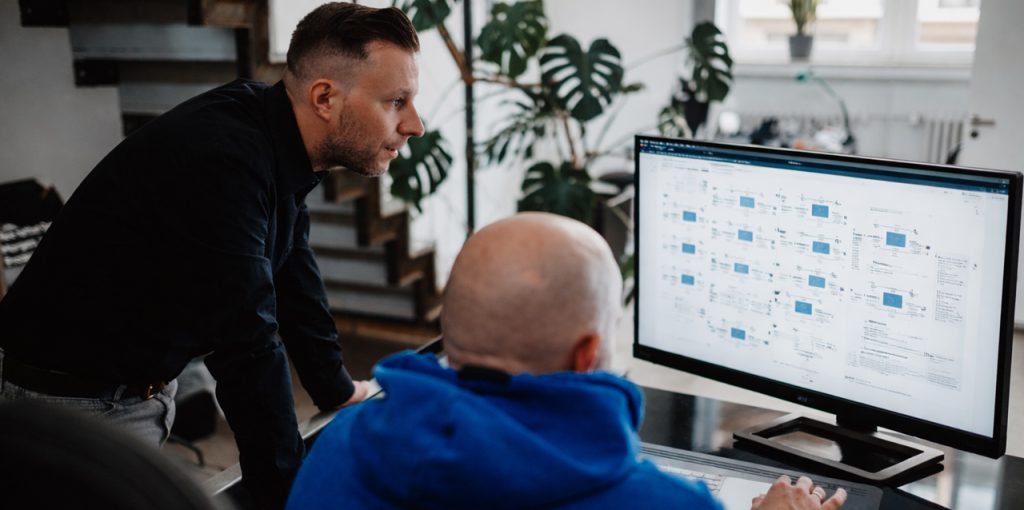 Zwei Männer analysieren gemeinsam einen technischen Ablauf an einem großen Monitor in einem modernen Büro. Der Bildschirm zeigt eine schematische Übersicht mit mehreren verbundenen Elementen, die an einen digitalen Prozess oder eine Systemarchitektur erinnert. Die Szene vermittelt Zusammenarbeit, Technikfokus und strukturierte Arbeit in ruhiger Atmosphäre.