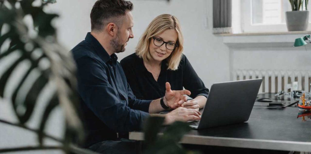 In einem hellen Büroraum sitzen ein Mann und eine Frau nebeneinander an einem dunklen Tisch. Beide blicken konzentriert auf einen geöffneten Laptop, der vor ihnen steht. Der Mann trägt ein dunkles Hemd und gestikuliert leicht mit der Hand, während er spricht. Die Frau mit schulterlangen blonden Haaren und Brille beugt sich leicht nach vorne und schaut aufmerksam auf den Bildschirm. Im Hintergrund sind ein Fenster mit Fensterbank, ein Heizkörper sowie eine Pflanze zu sehen. Eine weitere Pflanze ist unscharf im Vordergrund. Die Atmosphäre wirkt ruhig und professionell, als würden die beiden gemeinsam an einem digitalen Projekt arbeiten oder Inhalte besprechen.